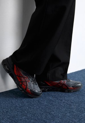 Zapatillas deportivas grises con detalles en rojo y patrón geométrico, con parte superior texturizada y suela de goma negra, colocadas sobre una alfombra azul.