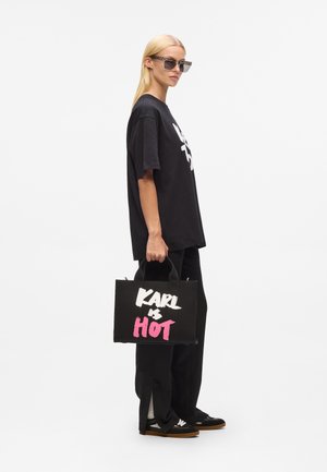 Sac fourre-tout noir avec "KARL & HOT" en texte blanc et rose, fabriqué en matériau résistant, avec des poignées courtes et une forme rectangulaire structurée.