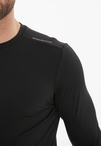 Sort langærmet atletisk top lavet af glat stof. Har en slim fit, blød tekstur og et logo "ENDURANCE" på skulderen.