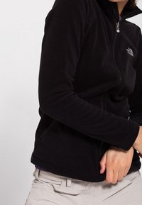 Musta fleece-takki korkealla kauluksella, vetskareilla ja huomaamattomalla logolla vasemmassa rinnassa. Varustettu sivutaskuilla ja pehmeällä tekstuurilla.