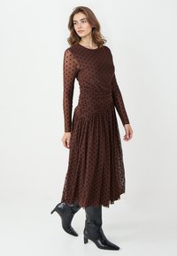 Bruine jurk met stippen, lange doorzichtige mouwen, geplooide taille, uitlopende rok en vloeiende stof. Gecombineerd met kniehoge zwarte puntige laarzen.