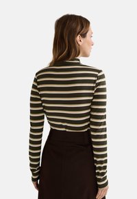 Haut à manches longues rayé en vert foncé avec des rayures beiges et blanches, en tissu lisse, au design col montant, associé à une jupe sombre taille haute.