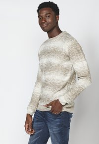 Beige en crème gemêleerde gebreide trui met een ronde hals, lange mouwen en geribbelde manchetten, gecombineerd met donkerblauwe denim jeans.