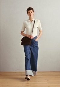 Weißes geripptes Polohemd, blaue weite Jeans mit weißem Saum, braune Umhängetasche und nacktfarbene Mules auf Holzboden vor einem hellen Hintergrund.