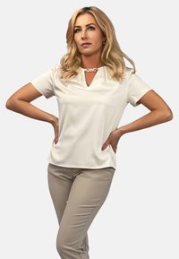 Krótki rękaw, ecru top z dekoltem w serek i dekoracyjnym złotym akcentem przy dekolcie, zestawiony z jasnobeżowymi dopasowanymi spodniami.