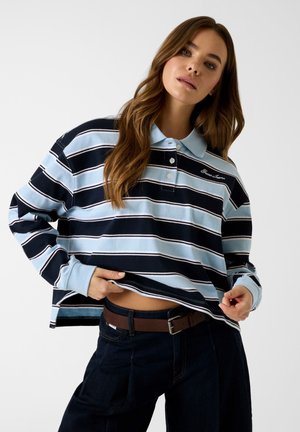 Femme portant un polo à manches longues à rayures bleu marine et bleu clair, un jean foncé et une ceinture marron, soulevant légèrement sa chemise pour dévoiler sa taille.