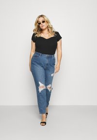 Zwarte geribbelde top met korte mouwen, hoog-waist lichtblauwe jeans met scheuren, en zwarte open teentjes hakken. Model draagt gele zonnebril.