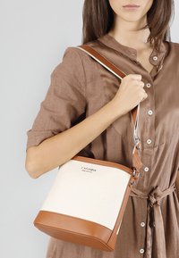 Bälte av tan läder och krämfärgad canvas med rektangulär form, silverfärgad hårdvara och justerbar rem. Har en logotyp på framsidan.