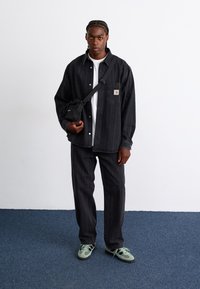 Schwarz gestreiftes Hemd und passende Hose, weißes T-Shirt mit Rundhalsausschnitt, schwarze Umhängetasche und grüne Sneakers. Einfaches Design, lässige Passform.