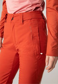 Pantalons de ski orange avec une texture lisse, taille ajustable et une poche latérale avec une fermeture éclair blanche. Les accents incluent un détail de logo.