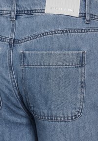 Vaaleansiniset farkut lähikuvassa, jossa on yksi takatasku, hienovaraisia ompelutaitoksia ja nahkainen brändilogo ommeltuna takataskun ylle.