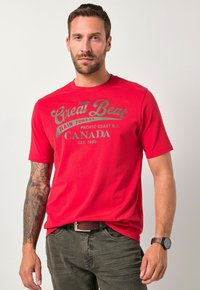Camiseta de algodón roja con estampado de "Great Bear Rain Forest" en verde y negro, escote redondo, mangas cortas, combinada con pantalones oliva y un cinturón marrón.