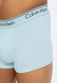 Άνδρας που φοράει ανοιχτό μπλε εσώρουχα boxer Calvin Klein, κοντινό πλάνο στη ζώνη και το πάνω μέρος του μηρού με λευκό φόντο.