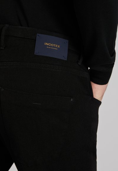 Close-up van de achterzak van een zwarte broek met een donkerblauw label met de tekst "INCOTEX BLUE DIVISION" en een hand die gedeeltelijk in de zak steekt.