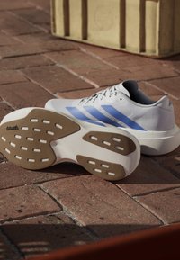 Scarpa da corsa bianca con strisce blu è sdraiata di lato su un pavimento in mattoni, mostrando la suola in gomma marrone con motivo antiscivolo e logo Continental.