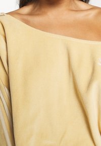 Gul velour-tröja med avskuren axel-design, med mjuk textur och en subtil broderad detalj på ena sidan.