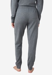 Persoon van achter in grijze gebreide broek en bijpassende trui, blootsvoets, staand tegen een eenvoudige witte achtergrond.