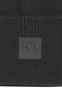 Mustassa neulepipoissa on ribboitu pinta ja neliön muotoinen musta nahkapaikka, jossa on kohokuvioitu "KARI TRAA" ja tyylitelty logo.