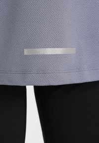 Ljuslila sporttopp med ett strukturerat, ventilerande tyg. Har en silverreflekterande remsa vid fållen för ökad synlighet.