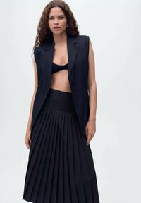 Gilet nero doppiopetto con spalle ampie, abbinato a una gonna plissè nera a vita alta. Entrambi i capi creano un look monocromatico.