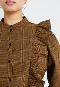 Camicia a quadri marrone con dettaglio arricciato sulla spalla, colletto alla coreana e bottoni neri lungo il davanti. Tessuto texture.