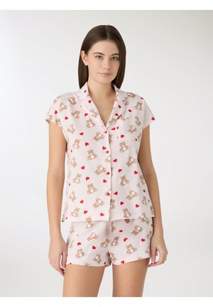 Giovane donna che indossa un pigiama rosa chiaro a maniche corte con motivi di orsacchiotti e cuori rossi, in piedi contro uno sfondo bianco semplice.