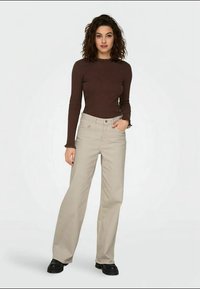 Brown long-sleeve top with frilled cuffs, paired with light beige wide-leg trousers and black chunky-soled shoes. Plain background.