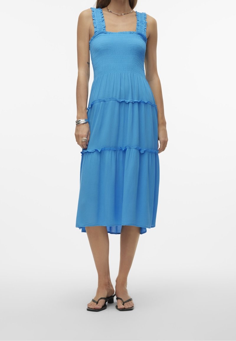 Femme portant une robe midi bleue sans manches à volants avec des bretelles à volants, associée à des sandales noire à talons et des bracelets argentés.