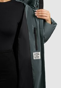 Giacca isolante verde con una tasca con zip e un'etichetta. L'imbottitura interna è nera, mostrando una finitura liscia e testurizzata.