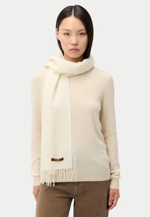 Frau trägt einen cremefarbenen Pullover und passenden Schal mit Fransen, kombiniert mit braunen Hosen, steht vor einem schlichten Hintergrund.