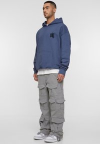 Blauer oversized Hoodie mit Fronttasche und schwarzem Logo, kombiniert mit grauen Cargo-Hosen mit mehreren Taschen und einem lockeren Schnitt. Weiße Sneakers.