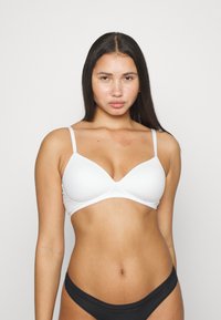 Bralette blanca acolchada con tirantes ajustables, textura suave y forma triangular clásica. Combinado con ropa interior negra.