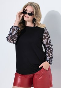 Czarny top z przezroczystymi, wzorzystymi, rozłożystymi rękawami; zestawiony z błyszczącymi czerwonymi szortami. Modelka poprawia czarne okulary przeciwsłoneczne i pokazuje kolczyki.