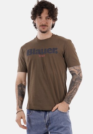 Uomo con capelli ricci che indossa una maglietta marrone "Blauer USA" e jeans blu, che mostra tatuaggi dettagliati in bianco e nero su entrambi gli avambracci.