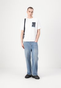 Camiseta blanca de manga corta con un bolsillo oscuro estampado, combinada con pantalones de mezclilla de pierna ancha en azul claro y zapatos negros con textura. Accesorio de correa negra.