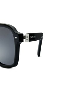 Zwarte zonnebril met een gedurfde rechthoekige montuur, grijze gradient lenzen en metalen accenten op de tempels met een ankerontwerp.