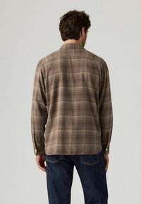 Uomo con camicia a quadri marrone a maniche lunghe e jeans blu scuro, in piedi di spalle contro uno sfondo chiaro uniforme.