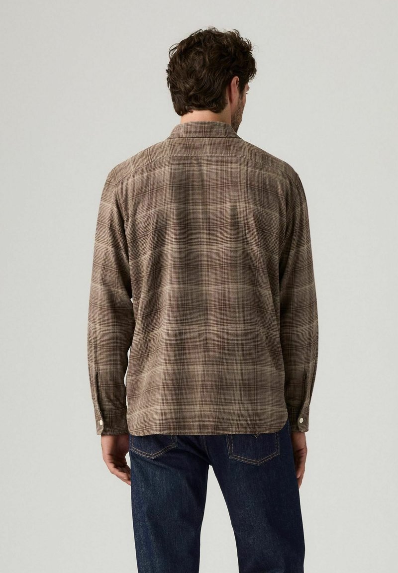 Mann, der ein braunes, kariertes Langarmhemd und dunkelblaue Jeans trägt, steht mit dem Rücken zur Kamera vor einem schlichten hellen Hintergrund.