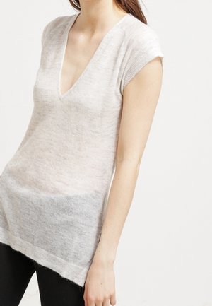 Femme portant un haut en maille beige clair à manches courtes avec col en V à bords côtelés, associé à un pantalon noir, sur un fond uni.
