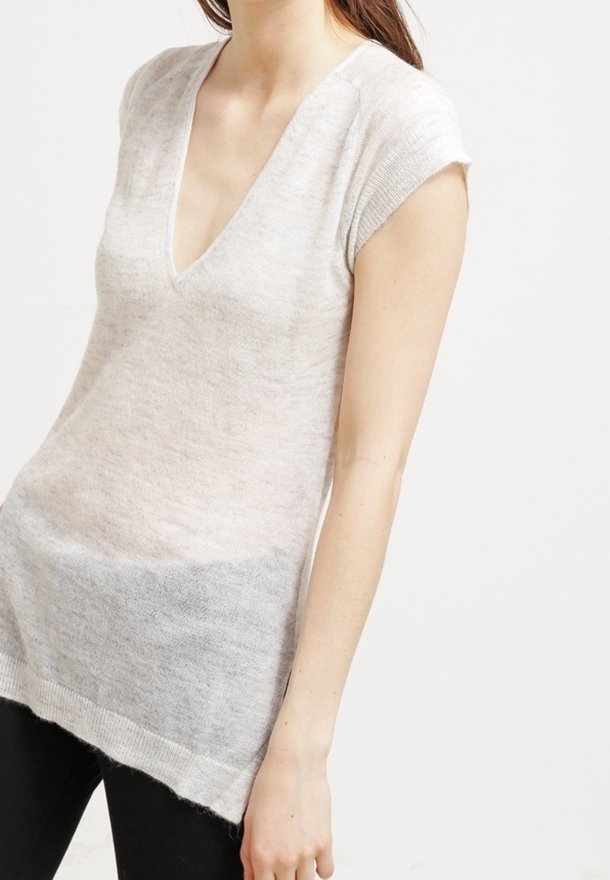 Femme portant un haut en maille beige clair à manches courtes avec col en V à bords côtelés, associé à un pantalon noir, sur un fond uni.