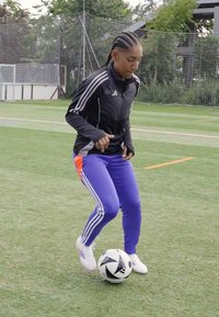 Persoon in een zwart Adidas-jack en paarse broek met oranje accenten dribbelt een zwart-wit voetbal op een grasveld.