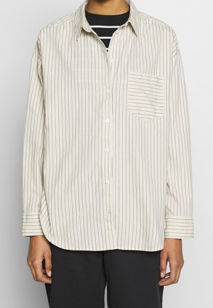 Personne portant une chemise beige ample à boutons avec de fines rayures verticales noires et une poche sur la poitrine, par-dessus un haut rayé noir et blanc.