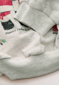 Hellgrauer Pullover mit Santa-Design und roten Akzenten. Mit gerippten Bündchen und weicher Textur. Enthält den Text "Happiest Festive Moments."