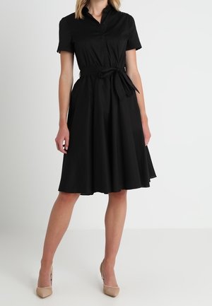 Kobieta w czarnej sukience z krótkim rękawem sięgającej do kolan, z kołnierzem i paskiem w talii, w beżowych szpilkach w szpic, stojąca na białej podłodze.