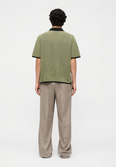 Άντρας με σγουρά μαλλιά που φοράει πράσινο πλεκτό πουλόβερ με κοντά μανίκια και φαρδιά μπεζ παντελόνια, στέκεται γυρισμένος με την πλάτη σε ένα απλό φόντο.