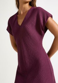 Kortærmet kjole i dyb lilla med V-udskæring og tekstureret stof med subtile vandrette striber. Slim-fit silhuet.