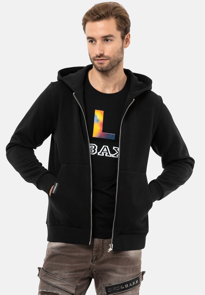 Hoodie noir zippé avec une poche frontale et un capuchon à cordon réglable, porté sur un t-shirt noir avec un design coloré.