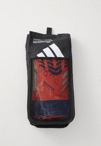 Guantes de portero de fútbol rojos en una bolsa de malla negra transparente. Presenta la marca Adidas, un asa superior y una sección inferior sólida de color azul.