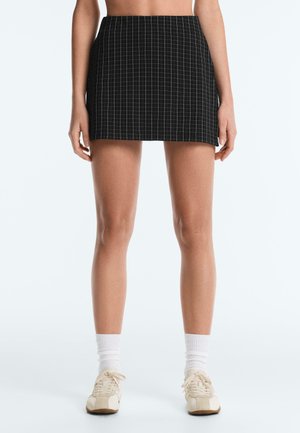 Kobieta w czarnej mini spódniczce z białym wzorem w kratę, białych skarpetkach i beżowych trampkach, stojąca na jednolitym białym tle.