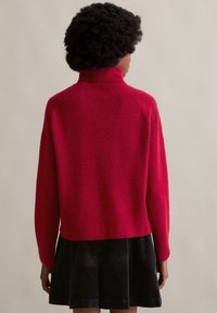 Roter Rollkragenpullover aus weichem, strukturiertem Strick. Hat ein verkürztes Design und einen gerippten Saum. Kombiniert mit einem schwarzen Cordrock.